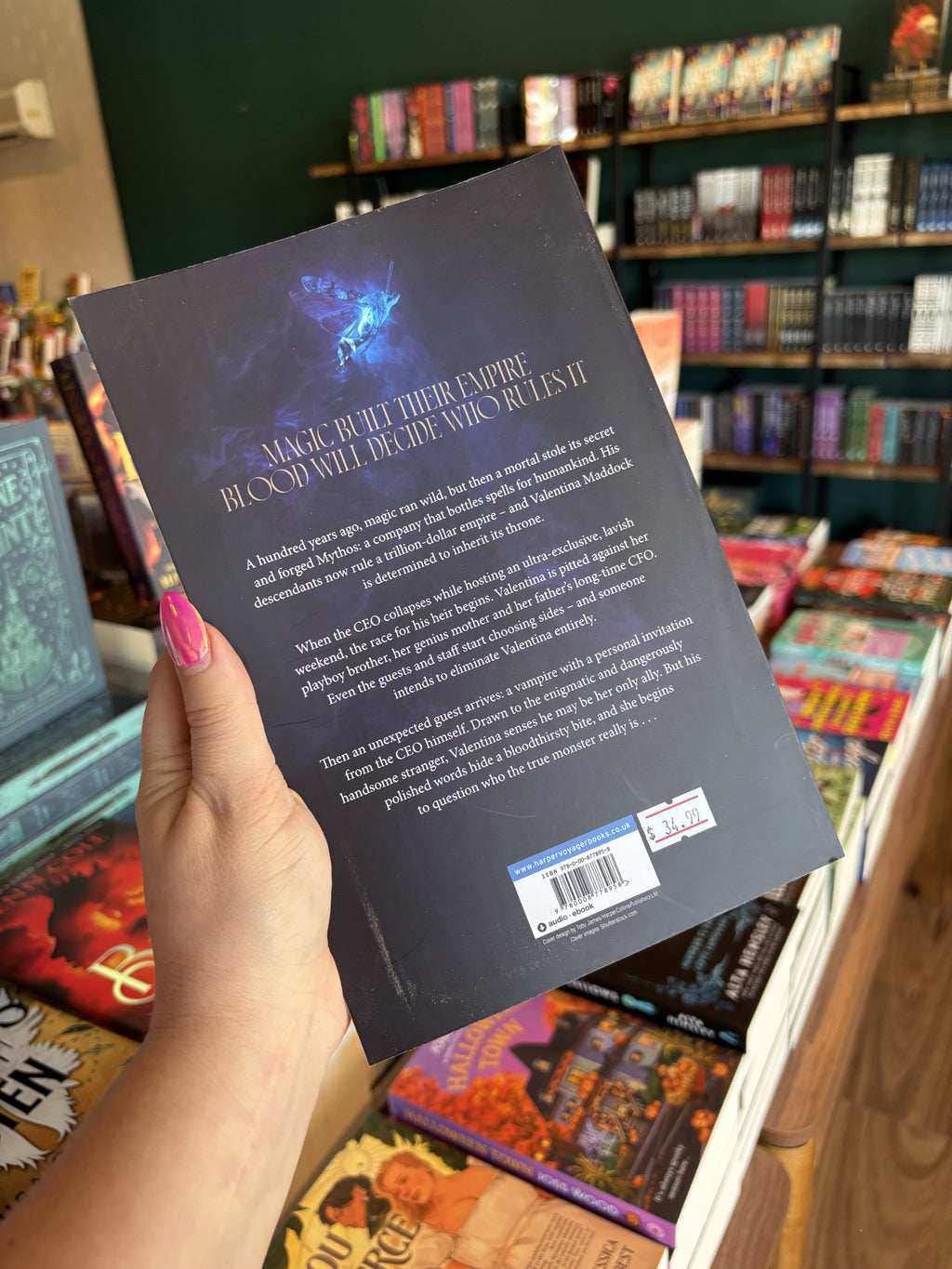 A hand with pink nail polish holding the back of a copy of 'The Age of Blood and Magic', featuring a dark background with a blue glowing moth, and light text.