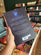 A hand with pink nail polish holding the back of a copy of 'The Age of Blood and Magic', featuring a dark background with a blue glowing moth, and light text.