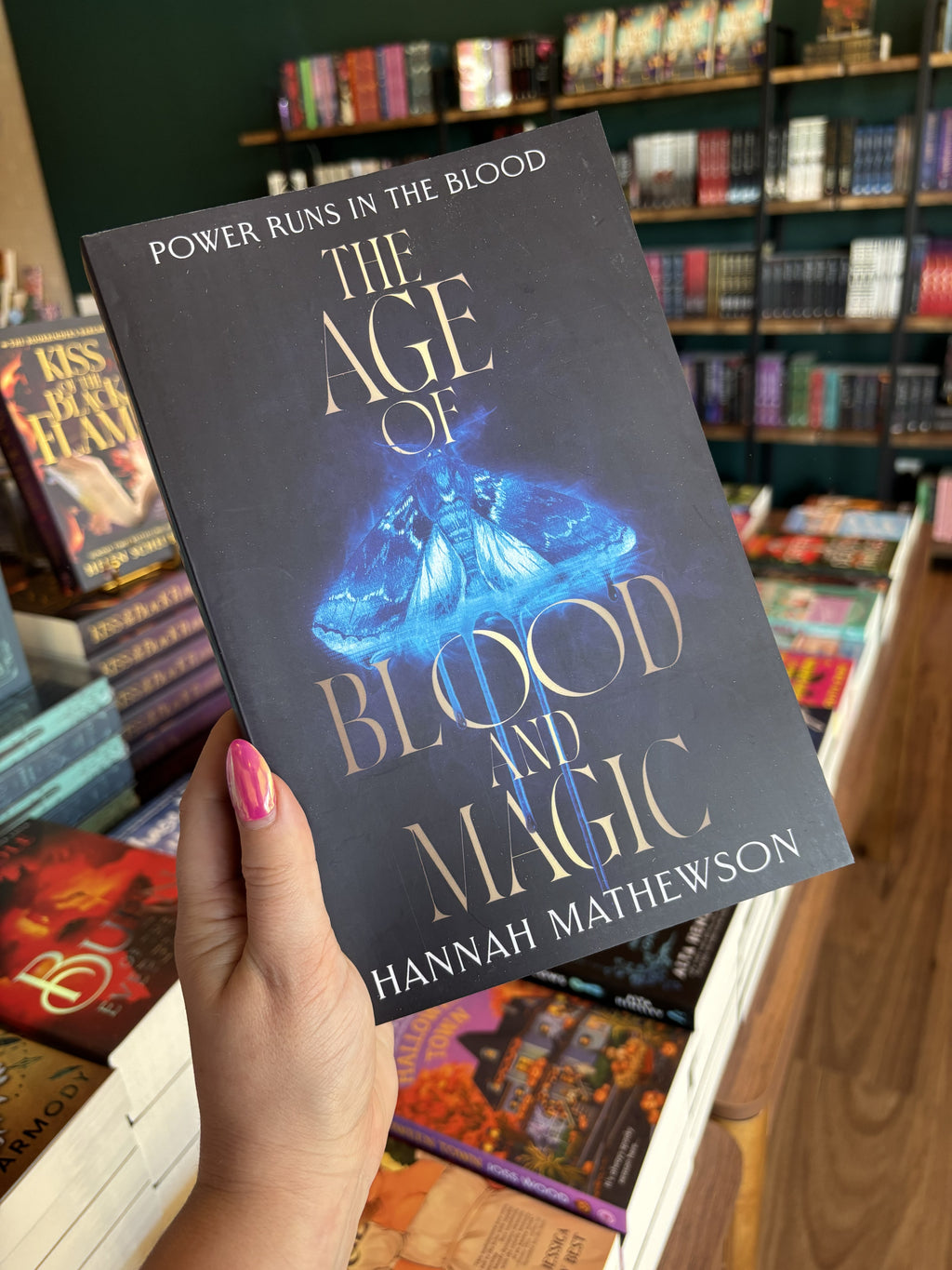 A hand with pink nail polish holding a copy of 'The Age of Blood and Magic', featuring a dark background with a blue glowing moth, and gold text.