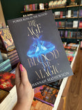 A hand with pink nail polish holding a copy of 'The Age of Blood and Magic', featuring a dark background with a blue glowing moth, and gold text.