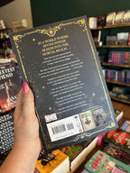 A hand with pink nail polish holding the back of a paperback copy of 'The Ascended' by Grenwich & Lennox, featuring a black background, a gold border, and white and gold text.