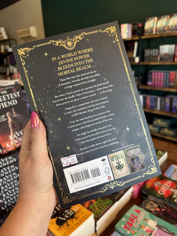 A hand with pink nail polish holding the back of a paperback copy of 'The Ascended' by Grenwich & Lennox, featuring a black background, a gold border, and white and gold text.