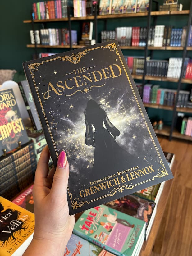 A hand with pink nail polish holding a paperback copy of 'The Ascended' by Grenwich & Lennox, featuring a dark silhouette of a woman in a flowing gown against a starry cosmic sky with ornate gold decorative borders and title text.