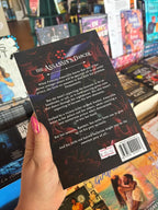A hand holds the book cover of 'The Assassin's Dancer' by Erin Fae Ryan featuring red lillies on a dark background with white font for the blurb, in front of a table full of other blurry books.