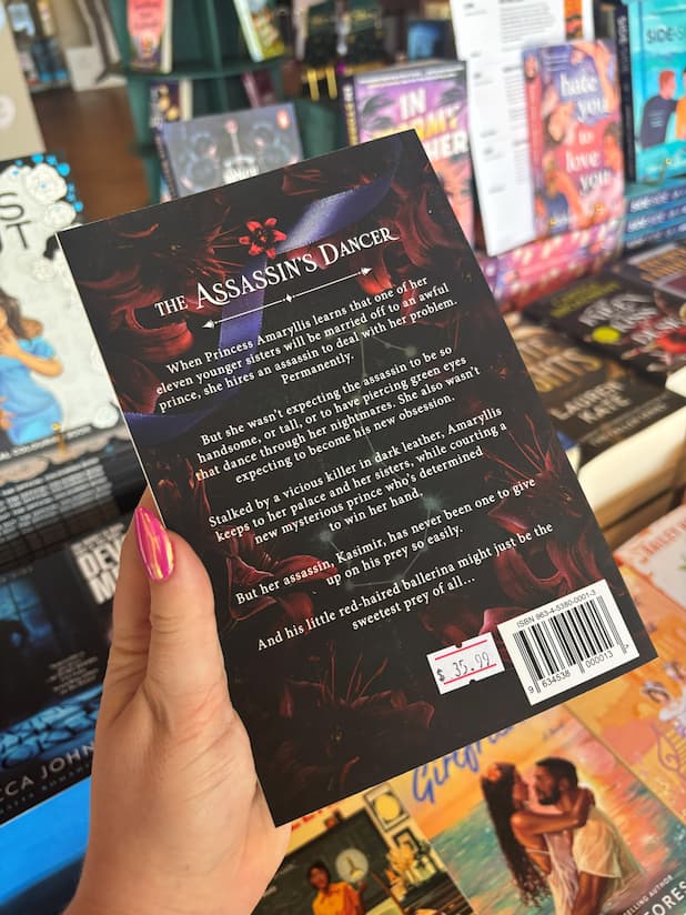 A hand holds the book cover of 'The Assassin's Dancer' by Erin Fae Ryan featuring red lillies on a dark background with white font for the blurb, in front of a table full of other blurry books.