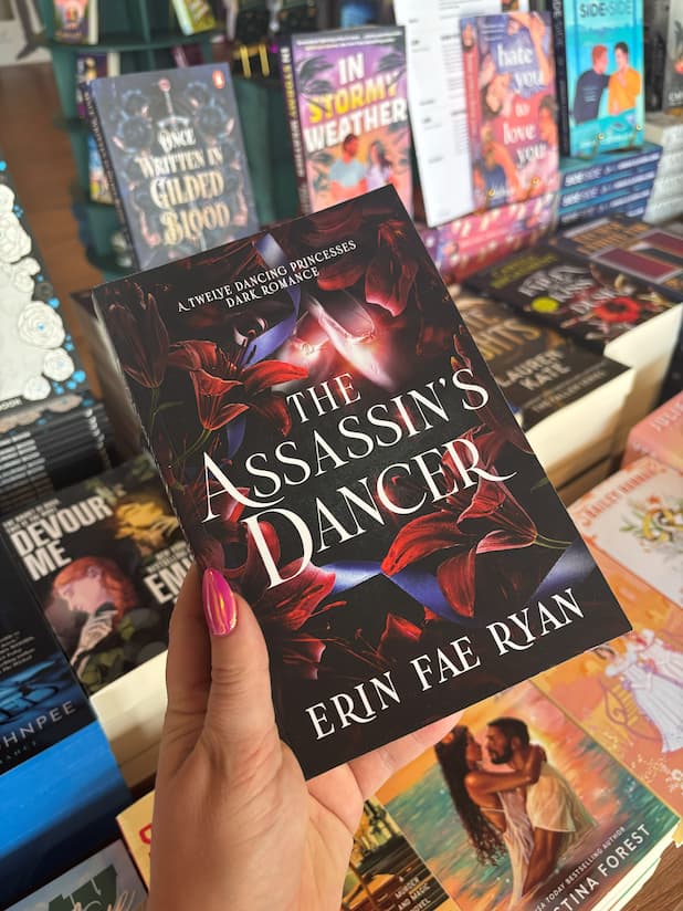 A hand holds the book cover of 'The Assassin's Dancer' by Erin Fae Ryan featuring red lillies on a dark background surrounding pink ballet slippers, in front of a table full of other blurry books.
