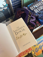 A hand holds the cover of 'The Assassin's Dancer' by Erin Fae Ryan open showing a gold signature under the title page, with an art print and matching bookmark poking out the top of the book, in front of a table full of other blurry books.