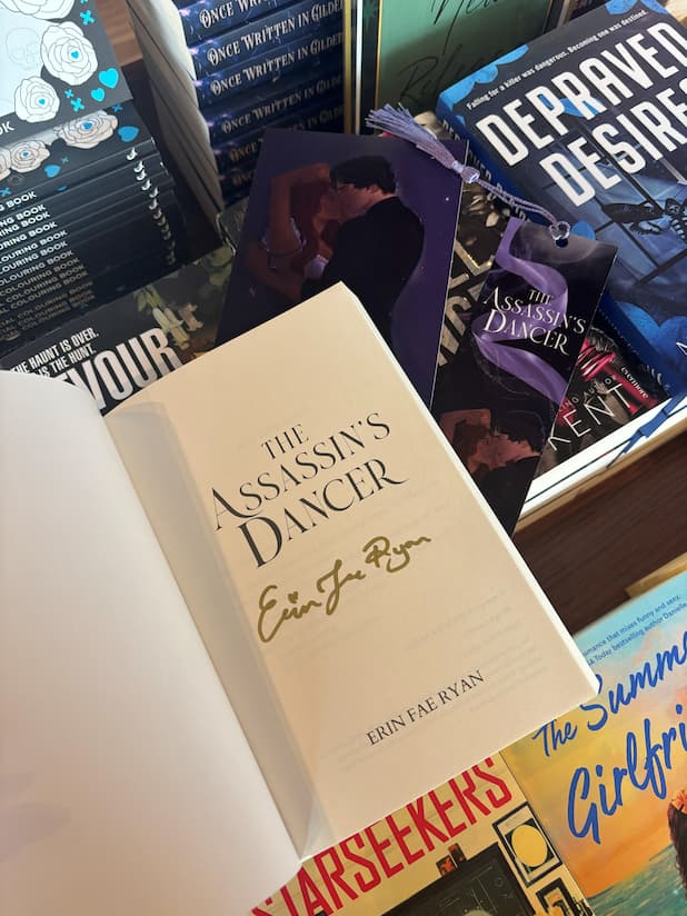 A hand holds the cover of 'The Assassin's Dancer' by Erin Fae Ryan open showing a gold signature under the title page, with an art print and matching bookmark poking out the top of the book, in front of a table full of other blurry books.