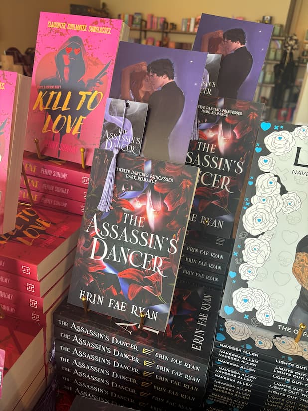 A stack of the book 'The Assassin's Dancer' by Erin Fae Ryan, with an art print and matching bookmark poking out the top of the book, featuring red lillies on a dark background surrounding pink ballet slippers, in front of a table full of other blurry books.