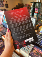 A hand holds the back cover for 'The Daddy's Girl' by Sara Cate, featuring a red to black gradient with white text for the blub. A red ribbon tied into a bow is in the bottom left corner of the cover.