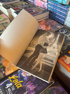 A hand holds open the cover for 'The Daddy's Girl' by Sara Cate, featuring character art of Amelia and Matis embraces in front of the eiffel tower at night, the book sits on a pile of other blurry books. 