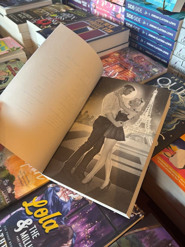 A hand holds open the cover for 'The Daddy's Girl' by Sara Cate, featuring character art of Amelia and Matis embraces in front of the eiffel tower at night, the book sits on a pile of other blurry books. 