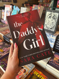 A hand holds the book cover for 'The Daddy's Girl' by Sara Cate, featuring large white title text over a red ribbon bow on a black and red background, in front of a blurry table full of books.