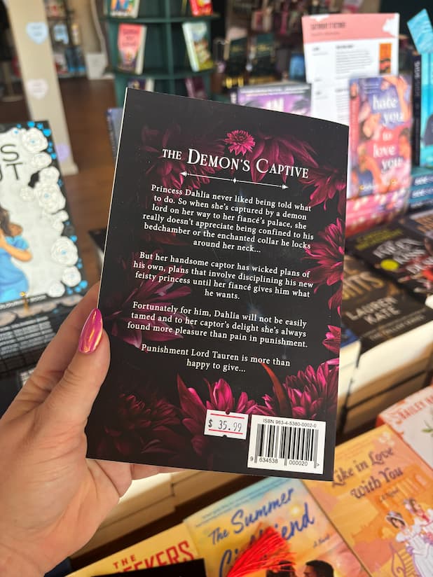 A hand holds the back cover of 'The Demon's Captive' by Erin Fae Ryan with dark magenta flowers framing the white text of the blurb, the book is held in front of a table full of other blurry books.