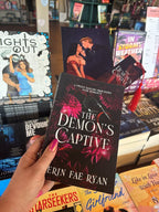 A hand holds the book cover of 'The Demon's Captive' by Erin Fae Ryan with dark magenta flowers and a whip, poking out the top is an art print and matching bookmark, the book is held in front of a table full of other blurry books.