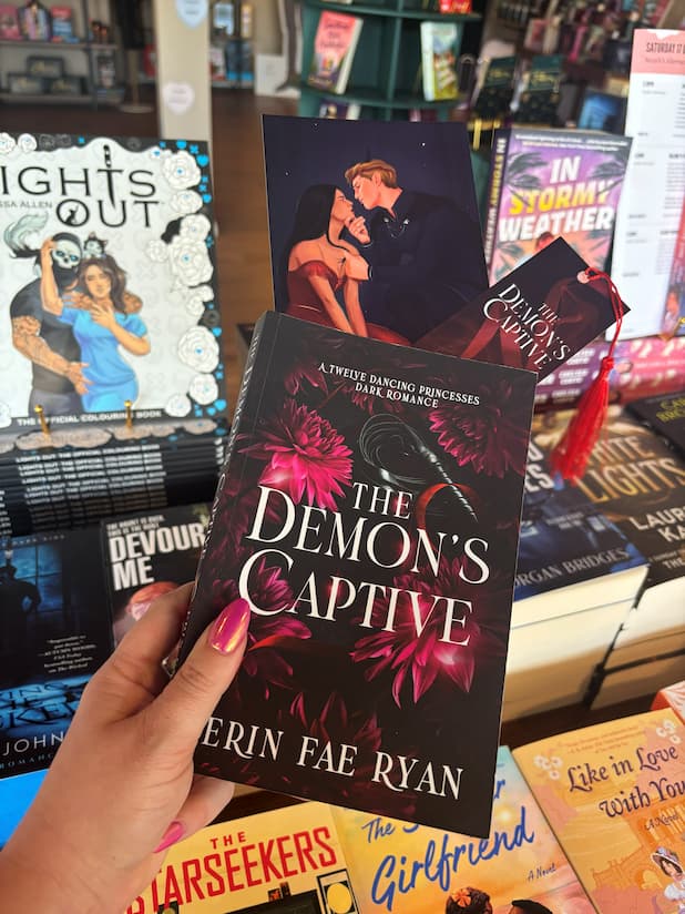 A hand holds the book cover of 'The Demon's Captive' by Erin Fae Ryan with dark magenta flowers and a whip, poking out the top is an art print and matching bookmark, the book is held in front of a table full of other blurry books.