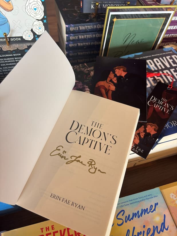 A hand holds an open copy of the book 'The Demon's Captive' by Erin Fae Ryan with a gold signature underneath the title on the first page, poking out the top is an art print and matching bookmark, the book is held in front of a table full of other blurry books.