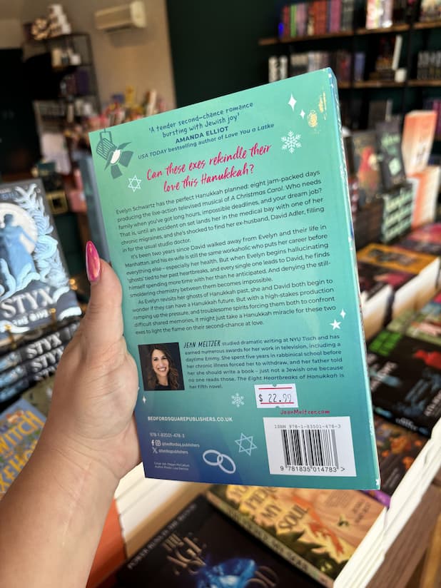 A hand with pink nail polish holding the back of a copy of 'The Eight Heartbreaks of Hanukkah' by Jean Meltzer, featuring a woman blue wintry background with pink and dark blue text.
