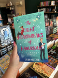A hand with pink nail polish holding a copy of 'The Eight Heartbreaks of Hanukkah' by Jean Meltzer, featuring a woman on her laptop and a man on his phone looking across the over at each other. The background is a gradient of blue and turquoise with Jewish icons sprinkled across the cover. 