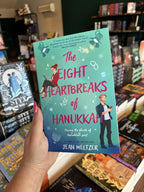 A hand with pink nail polish holding a copy of 'The Eight Heartbreaks of Hanukkah' by Jean Meltzer, featuring a woman on her laptop and a man on his phone looking across the over at each other. The background is a gradient of blue and turquoise with Jewish icons sprinkled across the cover. 