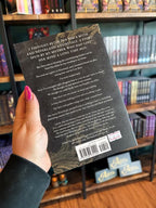 A hand with pink nail polish holds the back of a copy of 'The Fae Princes' by Nikki St. Crowe, featuring a textured back background with gold filigree on the corners, and white text.