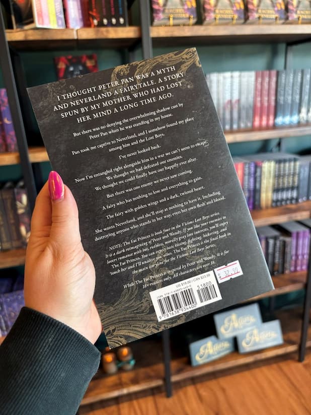 A hand with pink nail polish holds the back of a copy of 'The Fae Princes' by Nikki St. Crowe, featuring a textured back background with gold filigree on the corners, and white text.