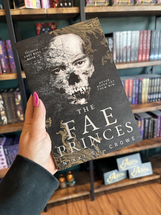A hand with pink nail polish holds a copy of 'The Fae Princes' by Nikki St. Crowe, featuring a textured back background with gold filigree on the corners, a disintegrating skull, and white text.