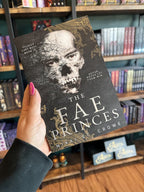 A hand with pink nail polish holds a copy of 'The Fae Princes' by Nikki St. Crowe, featuring a textured back background with gold filigree on the corners, a disintegrating skull, and white text.