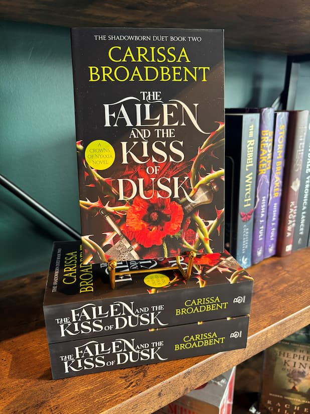 Three copies of the book 'The Fallen and the Kiss of Dusk' by Carissa Broadbent are stacked on a wooden bookshelf, with the top copy standing upright to show the front cover. The bookshelf is lined with other books, and a dark teal wall is visible in the background.