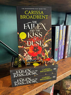 Three copies of the book 'The Fallen and the Kiss of Dusk' by Carissa Broadbent are stacked on a wooden bookshelf, with the top copy standing upright to show the front cover. The bookshelf is lined with other books, and a dark teal wall is visible in the background.