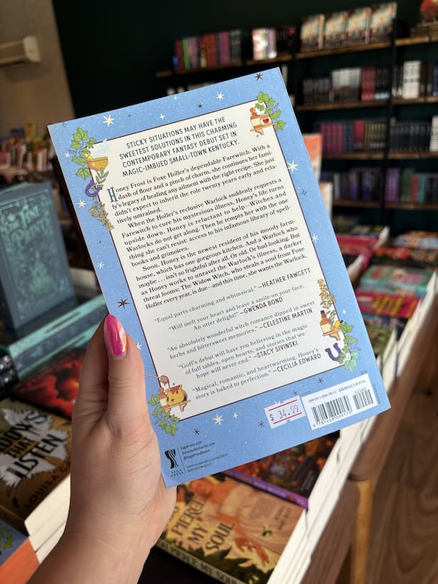 A hand with pink nail polish holding the back of a copy of 'The Farewitch of Foxholler' by Ellen Paley Goff, featuring whimsical fantasy artwork on a light blue background and dark text on a light section in the middle.
