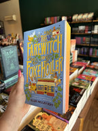 A hand with pink nail polish holding a copy of 'The Farewitch of Foxholler' by Ellen Paley Goff, featuring whimsical fantasy artwork on a light blue background with large yellow title text and illustrated characters, animals, and objects, and orange sprayed edges.