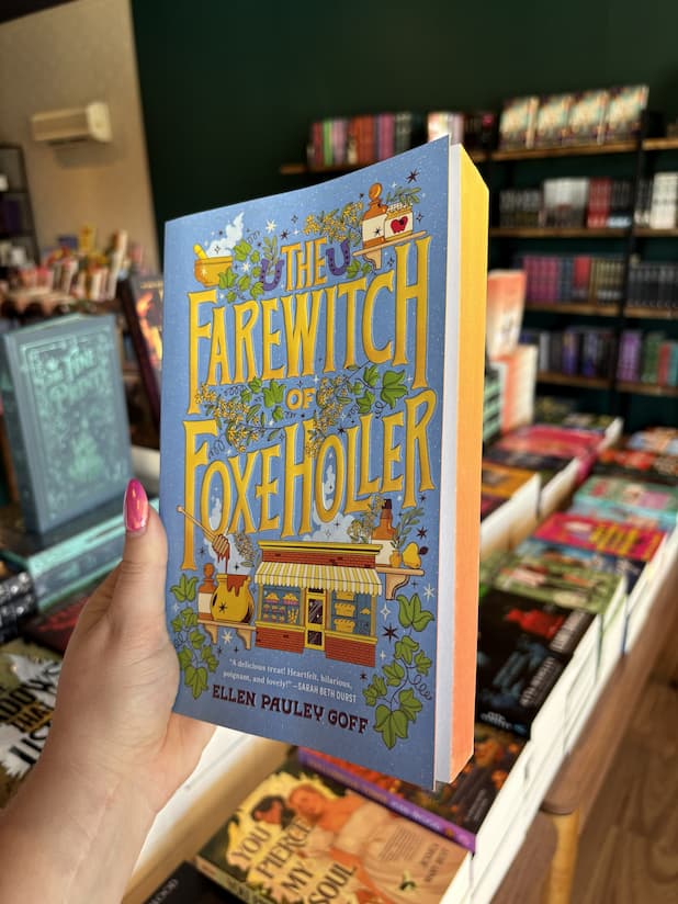 A hand with pink nail polish holding a copy of 'The Farewitch of Foxholler' by Ellen Paley Goff, featuring whimsical fantasy artwork on a light blue background with large yellow title text and illustrated characters, animals, and objects, and orange sprayed edges.