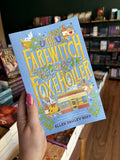A hand with pink nail polish holding a copy of 'The Farewitch of Foxholler' by Ellen Paley Goff, featuring whimsical fantasy artwork on a light blue background with large yellow title text and illustrated characters, animals, and objects.