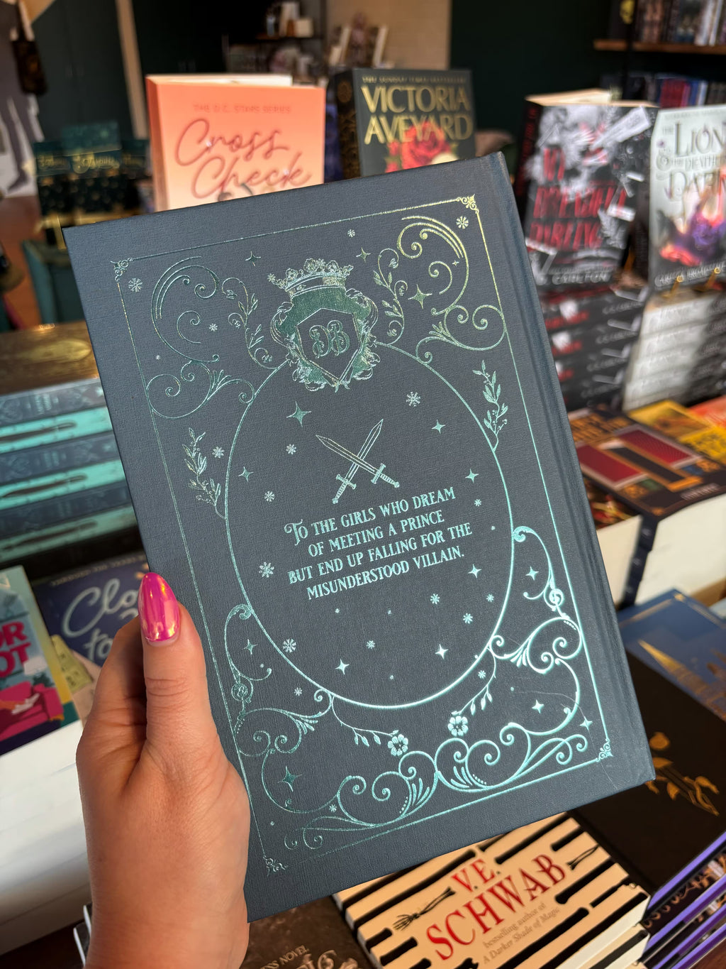 A hand with pink nail polish holds the back cover of the special edition version of 'The Fine Print' by Lauren Asher in a bookstore with other books blurry in the background. The cover is a dark teal with turquoise foiled decorative details around the dedication from the book.
