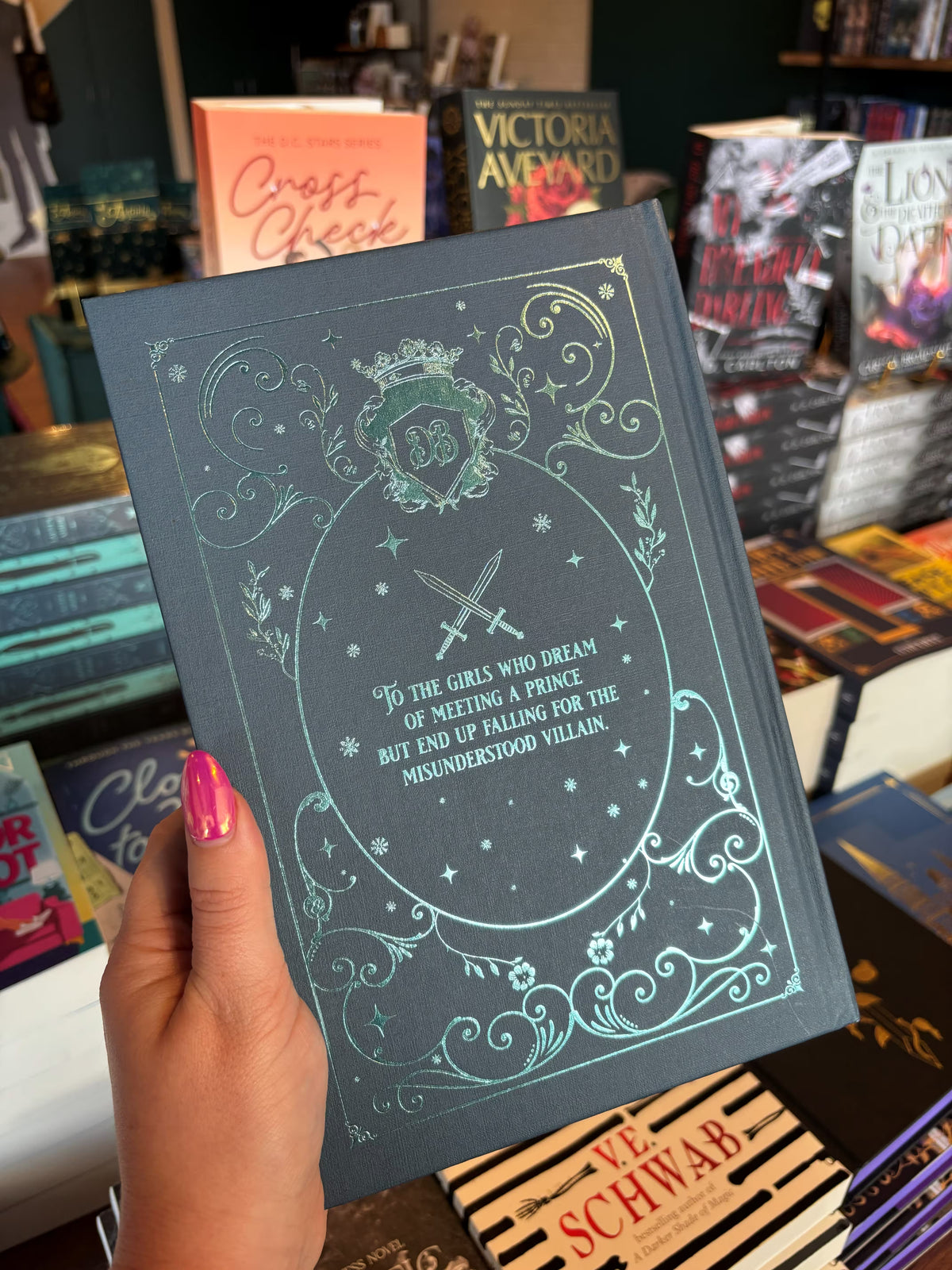 A hand with pink nail polish holds the back cover of the special edition version of 'The Fine Print' by Lauren Asher in a bookstore with other books blurry in the background. The cover is a dark teal with turquoise foiled decorative details around the dedication from the book.