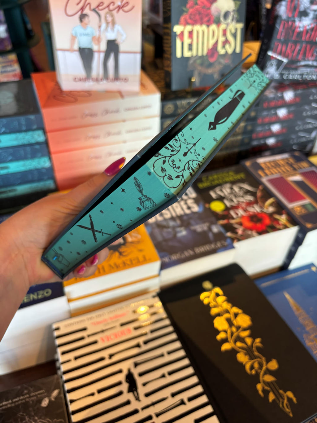 A hand with pink nail polish holds a copy of the special edition version of 'The Fine Print' by Lauren Asher in a bookstore with other books blurry in the background. The edges are an aqua colour with black illustrations of a pen and a sword surrounded by stars and filigree detailing. 