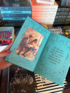 A hand with pink nail polish holds a copy of the special edition version of 'The Fine Print' by Lauren Asher in a bookstore with other books blurry in the background. The front end pages feature an illustration of the main characters sitting on a bench reading over pieces of paper on one side, and the book dedication in decorative text on the other side.