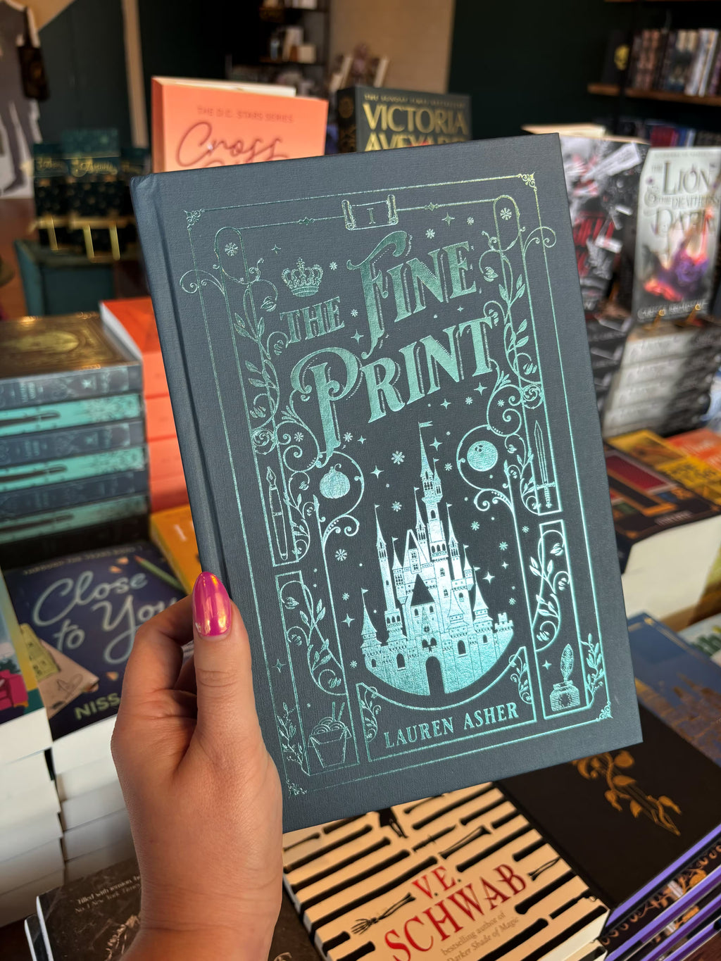 A hand with pink nail polish holds a copy of the special edition version of 'The Fine Print' by Lauren Asher in a bookstore with other books blurry in the background. The cover is a dark teal with turquoise foiled details of a castle, stars, and decorative borders.