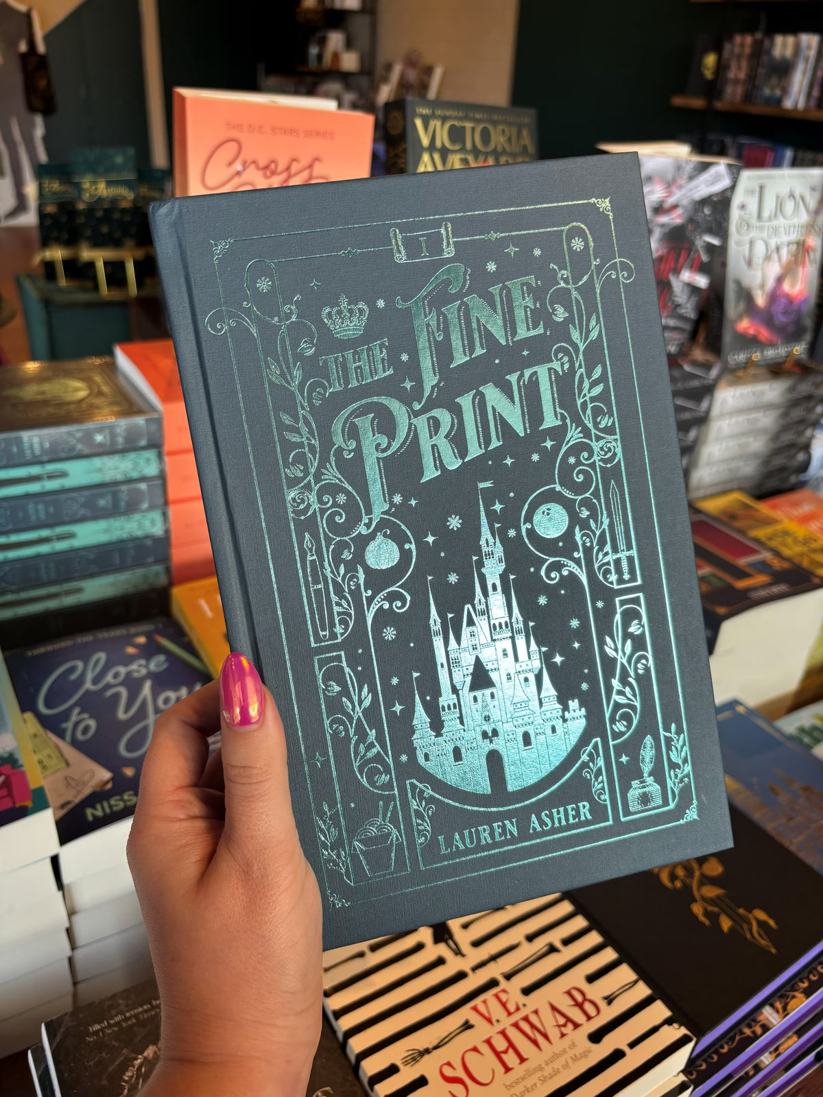 A hand with pink nail polish holds a copy of the special edition version of 'The Fine Print' by Lauren Asher in a bookstore with other books blurry in the background. The cover is a dark teal with turquoise foiled details of a castle, stars, and decorative borders.