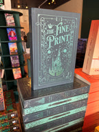 A stack of the special edition version of 'The Fine Print' by Lauren Asher in a bookstore with other books blurry in the background. The cover is a dark teal with turquoise foiled details of a castle, stars, and decorative borders.