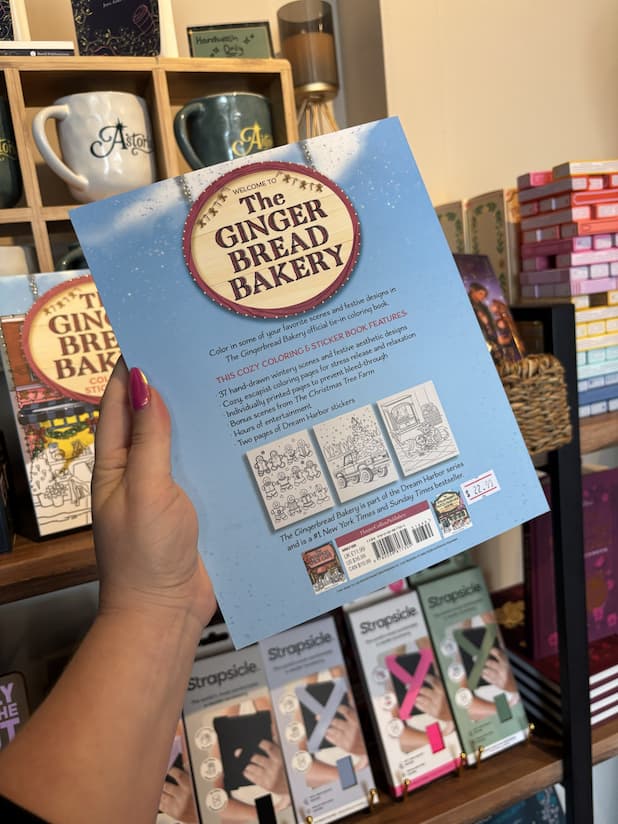A hand with pink nail polish holding the back of a copy of 'The Gingerbread Bakery Coloring & Sticker Book', featuring a sky blue background, black text, and sample colouring pages. 