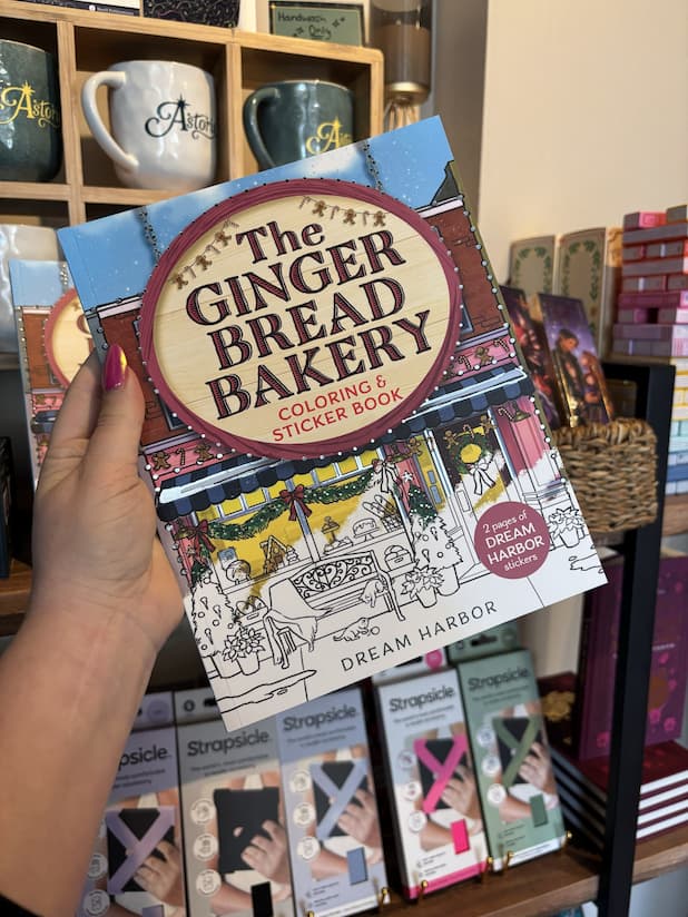 A hand with pink nail polish holding a copy of 'The Gingerbread Bakery Coloring & Sticker Book' with a festive bakery illustration half coloured in.