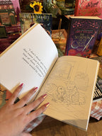 A hand with pink nail polish holding open a copy of 'The Gingerbread Bakery Coloring & Sticker Book' showing a quote on the left page and an illustration of two cats playing with a ball of wool.