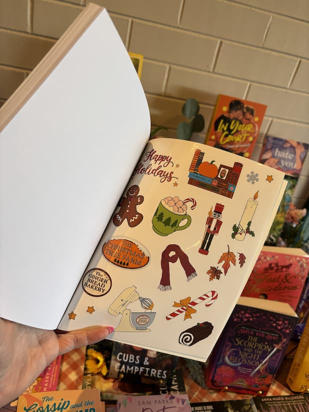 A hand with pink nail polish holding open a copy of 'The Gingerbread Bakery Coloring & Sticker Book' showing a page of festive stickers.