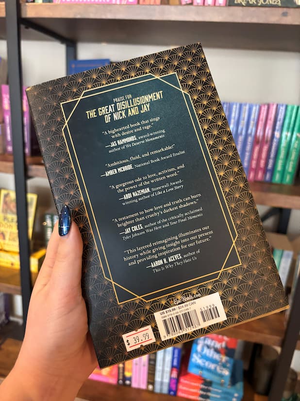 A hand holds the back cover of 'The Great Disillusionment of Nick and Jay' by Ryan Douglass with gold coloured font on a teal rectangle framed by gold art deco patterns on the cover.