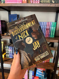 A hand holds the book cover of 'The Great Disillusionment of Nick and Jay' by Ryan Douglass in front of a blurry wooden bookshelf, with two men looking at each other from opposite corners of the cover against a cityscape on the top and a vintage club scene on the bottom.