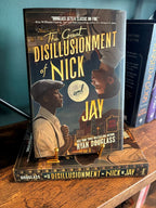 A copy of 'The Great Disillusionment of Nick and Jay' by Ryan Douglass sits on a  gold stand on top of another copy of the book on a wooden shelf. The cover features with two men looking at each other from opposite corners of the cover against a cityscape on the top and a vintage club scene on the bottom.