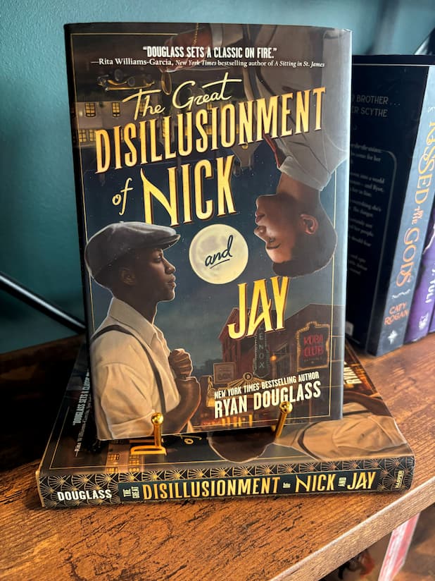 A copy of 'The Great Disillusionment of Nick and Jay' by Ryan Douglass sits on a  gold stand on top of another copy of the book on a wooden shelf. The cover features with two men looking at each other from opposite corners of the cover against a cityscape on the top and a vintage club scene on the bottom.
