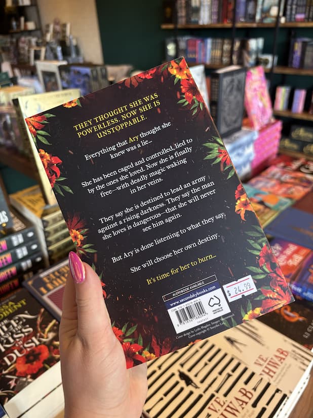 A hand with pink nails holding the back cover for 'The Incendiary' by LJ Claren, featuring white text against a dark background, framed by red and orange flowers.